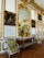 An opulent interior setting featuring ornate gold-trimmed walls, elaborate framed portraits, and a large decorative mirror. Between the portraits is an intricately carved wooden console topped with gilded decorations, and a pair of elegant green upholstered chairs flanking the console. The parquet wooden floor adds to the luxurious atmosphere.
