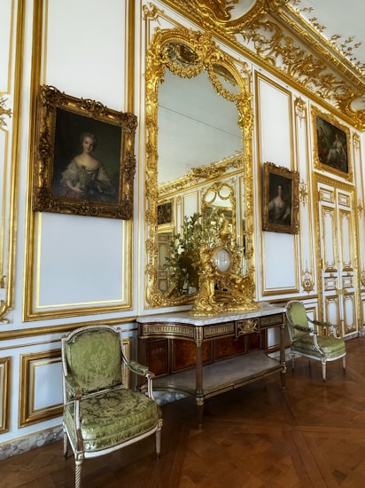 An opulent interior setting featuring ornate gold-trimmed walls, elaborate framed portraits, and a large decorative mirror. Between the portraits is an intricately carved wooden console topped with gilded decorations, and a pair of elegant green upholstered chairs flanking the console. The parquet wooden floor adds to the luxurious atmosphere.
