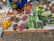 A market stall displays an assortment of fresh vegetables and fruits laid out on newspaper and cloth. The selection includes papayas, yams, sweet potatoes, cucumbers, green beans, peppers, and guavas. Brassware is arranged on one side. The person selling the goods is partially visible, wearing colorful traditional clothing.