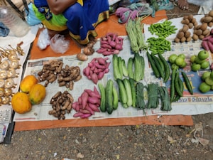 A market stall displays an assortment of fresh vegetables and fruits laid out on newspaper and cloth. The selection includes papayas, yams, sweet potatoes, cucumbers, green beans, peppers, and guavas. Brassware is arranged on one side. The person selling the goods is partially visible, wearing colorful traditional clothing.