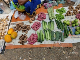 A market stall displays an assortment of fresh vegetables and fruits laid out on newspaper and cloth. The selection includes papayas, yams, sweet potatoes, cucumbers, green beans, peppers, and guavas. Brassware is arranged on one side. The person selling the goods is partially visible, wearing colorful traditional clothing.