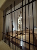 A wooden sculpture stands in the center of a dimly lit gallery space, viewed through vertical black bars resembling a gate or barrier. The room features polished wooden floors and multiple spotlights illuminating the artwork. The sculpture is abstract with layered elements and stands on a pedestal.