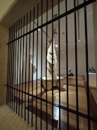 A wooden sculpture stands in the center of a dimly lit gallery space, viewed through vertical black bars resembling a gate or barrier. The room features polished wooden floors and multiple spotlights illuminating the artwork. The sculpture is abstract with layered elements and stands on a pedestal.