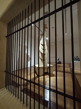 A wooden sculpture stands in the center of a dimly lit gallery space, viewed through vertical black bars resembling a gate or barrier. The room features polished wooden floors and multiple spotlights illuminating the artwork. The sculpture is abstract with layered elements and stands on a pedestal.