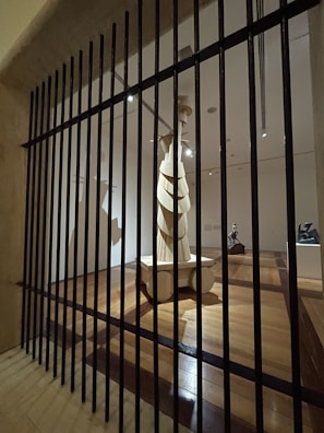 A wooden sculpture stands in the center of a dimly lit gallery space, viewed through vertical black bars resembling a gate or barrier. The room features polished wooden floors and multiple spotlights illuminating the artwork. The sculpture is abstract with layered elements and stands on a pedestal.