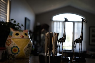 A kitchen countertop features a colorful ceramic owl-shaped container adorned with floral patterns. Beside it is a knife block holding several stainless steel knives. In the background, two decorative tall metal giraffe figurines stand before a window with sheer curtains that allow soft natural light to filter in.