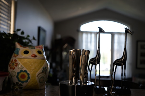 A kitchen countertop features a colorful ceramic owl-shaped container adorned with floral patterns. Beside it is a knife block holding several stainless steel knives. In the background, two decorative tall metal giraffe figurines stand before a window with sheer curtains that allow soft natural light to filter in.