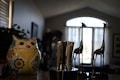 A kitchen countertop features a colorful ceramic owl-shaped container adorned with floral patterns. Beside it is a knife block holding several stainless steel knives. In the background, two decorative tall metal giraffe figurines stand before a window with sheer curtains that allow soft natural light to filter in.