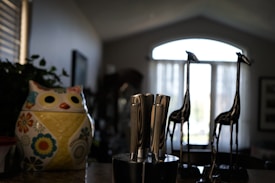 A kitchen countertop features a colorful ceramic owl-shaped container adorned with floral patterns. Beside it is a knife block holding several stainless steel knives. In the background, two decorative tall metal giraffe figurines stand before a window with sheer curtains that allow soft natural light to filter in.