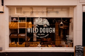 A bakery storefront with a large window displaying an assortment of bread and baked goods. The glass is adorned with the logo 'WILD DOUGH' along with the tagline 'REAL BREAD - REAL FOOD - LOCALLY SOURCED'. Inside, there are several shelves with loaves of bread and pastries. The atmosphere suggests a welcoming and artisanal vibe, further highlighted by warm lighting from hanging lamps.