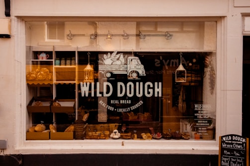 A bakery storefront with a large window displaying an assortment of bread and baked goods. The glass is adorned with the logo 'WILD DOUGH' along with the tagline 'REAL BREAD - REAL FOOD - LOCALLY SOURCED'. Inside, there are several shelves with loaves of bread and pastries. The atmosphere suggests a welcoming and artisanal vibe, further highlighted by warm lighting from hanging lamps.