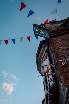 A pub sign labeled 'The Bull Brakspear' hangs from a brick building with a bay window. Red, white, and blue triangular flags are strung across the sky, suggesting a festive atmosphere. The sky is clear with a few clouds, and warm light emanates from the window.