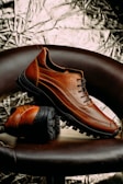 A pair of brown leather shoes with black rugged soles is artfully displayed. One shoe is upright, resting partially on the sole of the other, which is lying flat. The background features a textured, metallic appearance with crumpled patterns. The chair they are placed on is dark brown, complementing the rich color of the shoes.