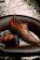 A pair of brown leather shoes with black rugged soles is artfully displayed. One shoe is upright, resting partially on the sole of the other, which is lying flat. The background features a textured, metallic appearance with crumpled patterns. The chair they are placed on is dark brown, complementing the rich color of the shoes.
