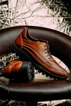 A pair of brown leather shoes with black rugged soles is artfully displayed. One shoe is upright, resting partially on the sole of the other, which is lying flat. The background features a textured, metallic appearance with crumpled patterns. The chair they are placed on is dark brown, complementing the rich color of the shoes.