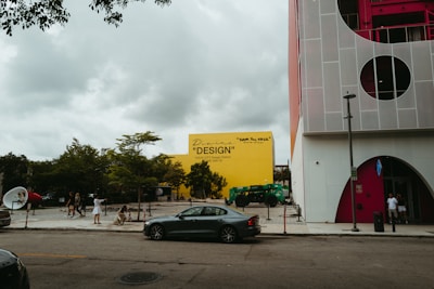 An urban street scene with a prominent yellow building in the background featuring text related to design. There are trees and greenery in front of the building. On the right, a modern structure is visible with circular windows and red accents. Pedestrians and a parked black car are present on the street, and the sky is overcast with clouds.