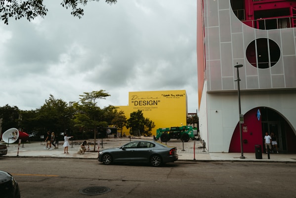 An urban street scene with a prominent yellow building in the background featuring text related to design. There are trees and greenery in front of the building. On the right, a modern structure is visible with circular windows and red accents. Pedestrians and a parked black car are present on the street, and the sky is overcast with clouds.