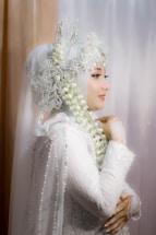 A woman is adorned in an elaborate white lace garment that includes a hijab integrated with ornate lace and beadwork details. Her profile view reveals a delicate arrangement of jasmine flowers entwined in her headdress. She exudes elegance and grace, with intricate lace gloves accentuating her hands.