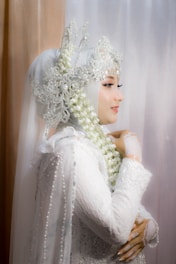 A woman is adorned in an elaborate white lace garment that includes a hijab integrated with ornate lace and beadwork details. Her profile view reveals a delicate arrangement of jasmine flowers entwined in her headdress. She exudes elegance and grace, with intricate lace gloves accentuating her hands.