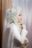 A woman is adorned in an elaborate white lace garment that includes a hijab integrated with ornate lace and beadwork details. Her profile view reveals a delicate arrangement of jasmine flowers entwined in her headdress. She exudes elegance and grace, with intricate lace gloves accentuating her hands.