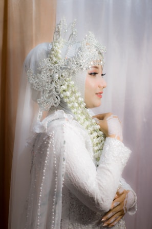A woman is adorned in an elaborate white lace garment that includes a hijab integrated with ornate lace and beadwork details. Her profile view reveals a delicate arrangement of jasmine flowers entwined in her headdress. She exudes elegance and grace, with intricate lace gloves accentuating her hands.