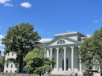 A classical-style building with tall white columns and a triangular pediment. The structure is surrounded by lush green trees, under a clear blue sky with a few clouds.