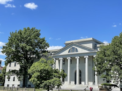 A classical-style building with tall white columns and a triangular pediment. The structure is surrounded by lush green trees, under a clear blue sky with a few clouds.