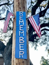 A wooden pole has a blue sign attached to it with the word 'REMEMBER' written vertically in white letters. Two American flags are placed on either side of the sign, and the scene is set outdoors with tree branches in the background.