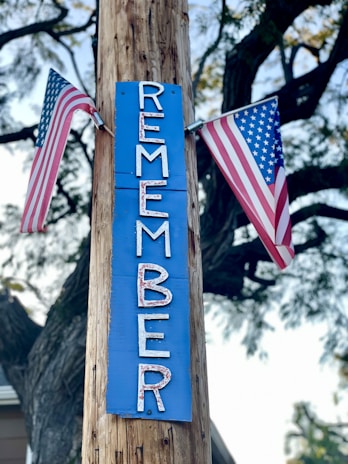 A wooden pole has a blue sign attached to it with the word 'REMEMBER' written vertically in white letters. Two American flags are placed on either side of the sign, and the scene is set outdoors with tree branches in the background.