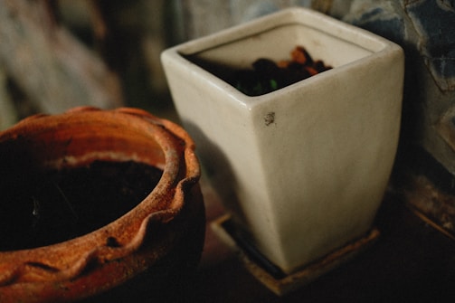 Two plant pots are in focus. One is a rustic, round terracotta pot, displaying a warm orange-brown hue. Next to it is a square, off-white ceramic pot, both containing dark soil inside. The background is slightly blurred, giving a cozy, earthy feel.