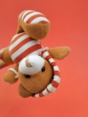 A plush toy resembling an animal is being held upside down against a solid red background. The toy is light brown with a white and red striped outfit, including a matching cap. Its black button-like eyes and round nose give it a cute, playful appearance.