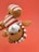 A plush toy resembling an animal is being held upside down against a solid red background. The toy is light brown with a white and red striped outfit, including a matching cap. Its black button-like eyes and round nose give it a cute, playful appearance.