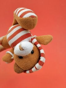 A plush toy resembling an animal is being held upside down against a solid red background. The toy is light brown with a white and red striped outfit, including a matching cap. Its black button-like eyes and round nose give it a cute, playful appearance.