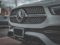 A close-up view of the front grill of a luxury car featuring a shiny emblem and intricate design details. The car's metallic finish reflects the surrounding environment, giving a sleek and polished appearance.