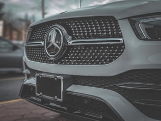 A close-up view of the front grill of a luxury car featuring a shiny emblem and intricate design details. The car's metallic finish reflects the surrounding environment, giving a sleek and polished appearance.