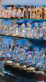 An array of ornate perfume bottles displayed on glass shelves. The bottles are crafted from crystal and various metals, featuring intricate designs with reflective surfaces. The arrangement highlights a variety of shapes and sizes, with the top shelves showcasing taller, elegant bottles and the bottom ones holding shorter jars with detailed caps.