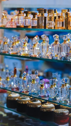 An array of ornate perfume bottles displayed on glass shelves. The bottles are crafted from crystal and various metals, featuring intricate designs with reflective surfaces. The arrangement highlights a variety of shapes and sizes, with the top shelves showcasing taller, elegant bottles and the bottom ones holding shorter jars with detailed caps.
