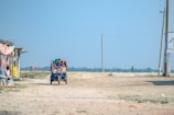 A three-wheeled vehicle carrying several people drives along a dirt path. There are makeshift structures to the side and a person walking in the distance. The sky is clear with minimal clouds, providing a bright and open atmosphere.