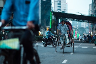 A busy urban street scene with several rickshaws and motorcycles. A man pedals a rickshaw in the center while others are around, including a person on a bicycle in the foreground. Tall buildings and an overhead passageway are visible in the background, with colorful lights lining the sides of the street.