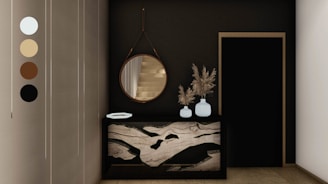 An elegant and modern interior space featuring a dark wall with a round mirror hanging above a decorative console table. The table has a unique marble design and holds two white vases filled with dried pampas grass. Next to it is a black door with a simple frame. Neutral tones are present, accentuated by the sleek design elements.