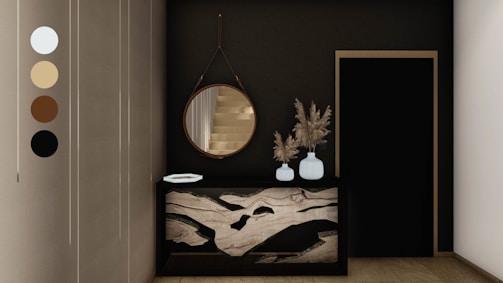 An elegant and modern interior space featuring a dark wall with a round mirror hanging above a decorative console table. The table has a unique marble design and holds two white vases filled with dried pampas grass. Next to it is a black door with a simple frame. Neutral tones are present, accentuated by the sleek design elements.