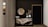An elegant and modern interior space featuring a dark wall with a round mirror hanging above a decorative console table. The table has a unique marble design and holds two white vases filled with dried pampas grass. Next to it is a black door with a simple frame. Neutral tones are present, accentuated by the sleek design elements.
