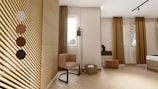 A modern interior space featuring a wooden-slatted partition with a color palette on the left side. Neutral tones are predominant, with light wood flooring and a beige accent chair. There are large windows with brown curtains and a floor lamp beside the chair. A small black side table holds a decorative piece, and an ottoman is placed nearby, along with a woven basket containing a rolled towel. The overall design emphasizes minimalism and natural elements.