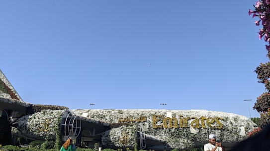 A large structure resembling an airplane is covered entirely in greenery and white flowers, with the word 'Emirates' prominently displayed in golden letters. The sky is clear and blue, with a few individuals visible in the foreground, one of whom is taking a photo.