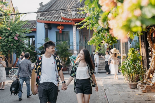 A smiling couple holding hands walks down a sunlit street lined with greenery and traditional architecture. Other people casually stroll in the background, creating a lively and warm atmosphere.