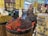 A market stall displays a variety of fruits in large round containers. Among them are strawberries, cherries, and grapes. The fruits are organized neatly, with some labels indicating their types and prices, such as Greek strawberries and sweet lychees. The vibrant colors of the fruits stand out against the retail environment. In the background, additional fruit and produce sections with people shopping are visible.