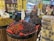 A market stall displays a variety of fruits in large round containers. Among them are strawberries, cherries, and grapes. The fruits are organized neatly, with some labels indicating their types and prices, such as Greek strawberries and sweet lychees. The vibrant colors of the fruits stand out against the retail environment. In the background, additional fruit and produce sections with people shopping are visible.