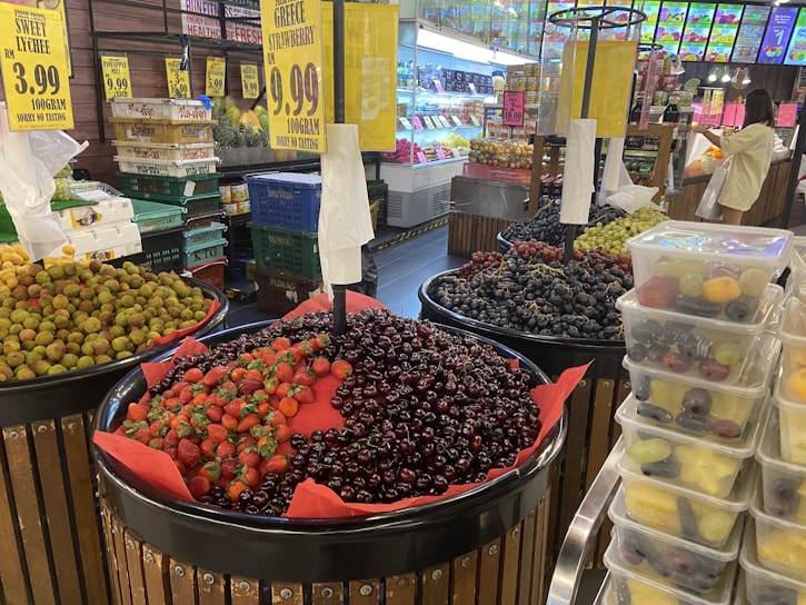 A market stall displays a variety of fruits in large round containers. Among them are strawberries, cherries, and grapes. The fruits are organized neatly, with some labels indicating their types and prices, such as Greek strawberries and sweet lychees. The vibrant colors of the fruits stand out against the retail environment. In the background, additional fruit and produce sections with people shopping are visible.