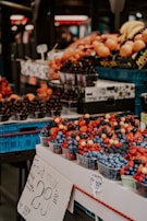 A market stall displays a variety of fresh fruits arranged in plastic containers. The fruits include strawberries, blueberries, blackberries, raspberries, and currants. A prominent sign indicates the price for a fruit mix. In the background, more fruits like bananas and oranges are visible, stacked in blue crates.
