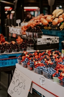 A market stall displays a variety of fresh fruits arranged in plastic containers. The fruits include strawberries, blueberries, blackberries, raspberries, and currants. A prominent sign indicates the price for a fruit mix. In the background, more fruits like bananas and oranges are visible, stacked in blue crates.